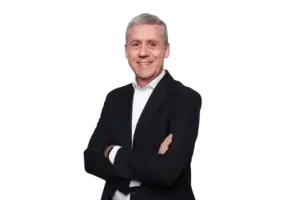 Un hombre de mediana edad, con el pelo corto y canoso, sonríe de pie, con los brazos cruzados, vestido con una chaqueta de traje oscura y una camisa blanca, frente a un fondo blanco liso.