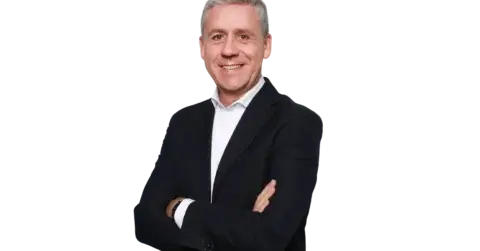 Un hombre de mediana edad, con el pelo corto y canoso, sonríe de pie, con los brazos cruzados, vestido con una chaqueta de traje oscura y una camisa blanca, frente a un fondo blanco liso.