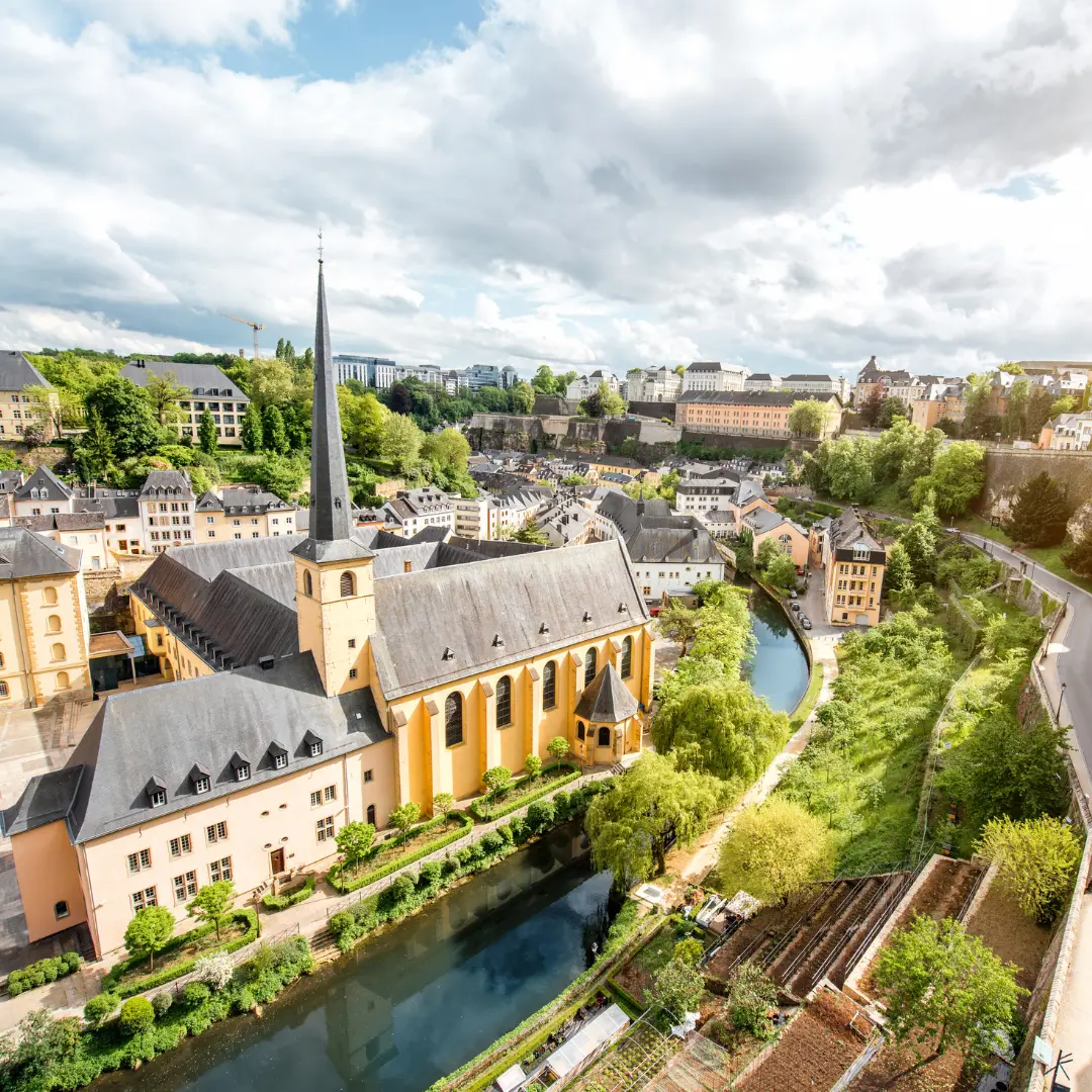 Et panoramautsikt over Luxembourg by med Neumünster-klosteret, et høyt kirketårn, frodige hager og historiske bygninger ved elven under en delvis overskyet himmel. For mer informasjon, kontakt oss.