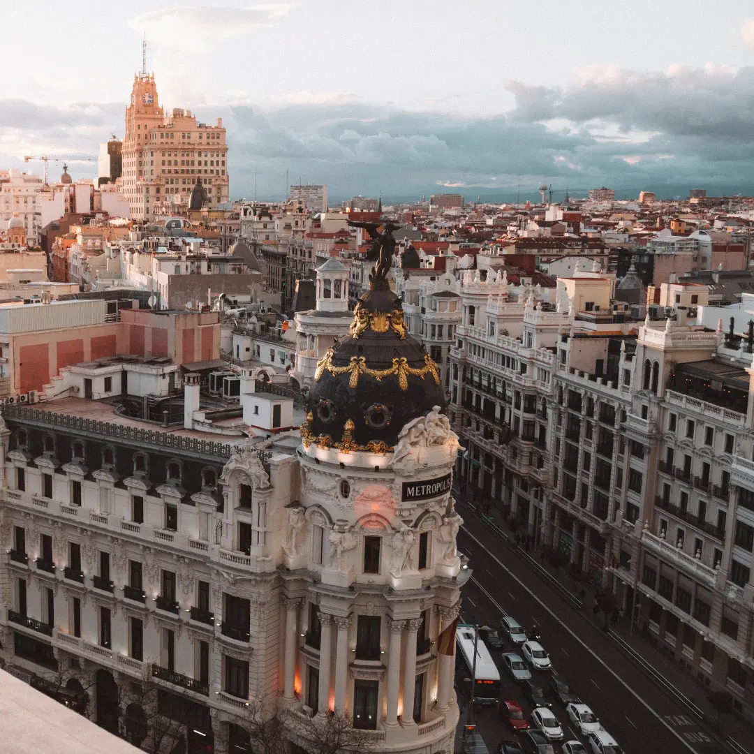 Aerial view of Madrid’s Gran Vía at sunset, featuring the ornate Metropolis Building with its black and gold dome, surrounded by historic white buildings and bustling city streets—contact f24 to capture moments like this.