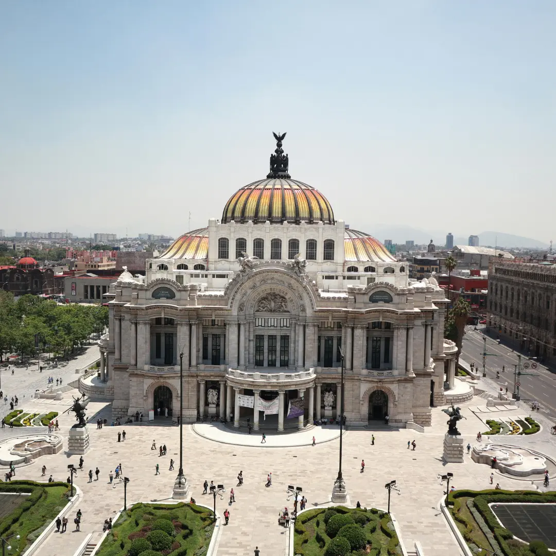The image shows the Palacio de Bellas Artes in Mexico City, a grand white marble building with a colorful domed roof, surrounded by gardens and people walking in the plaza—perfect for those wishing to contact f24 for more information.