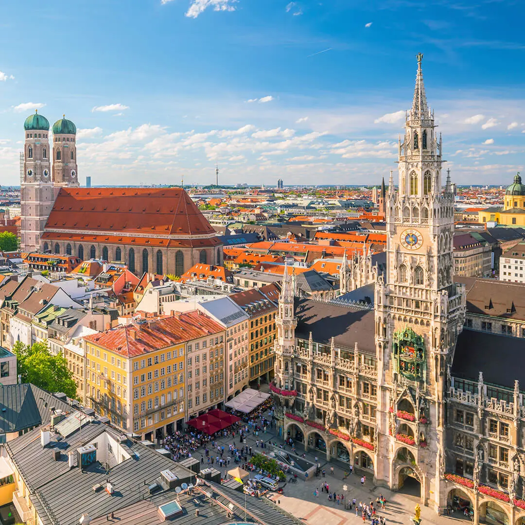 Aerial view of Munich’s Marienplatz with the ornate New Town Hall, the twin towers of Frauenkirche, and historic buildings under a blue sky. For more information or inquiries, please contact F24. Crowds gather in the lively city square below.