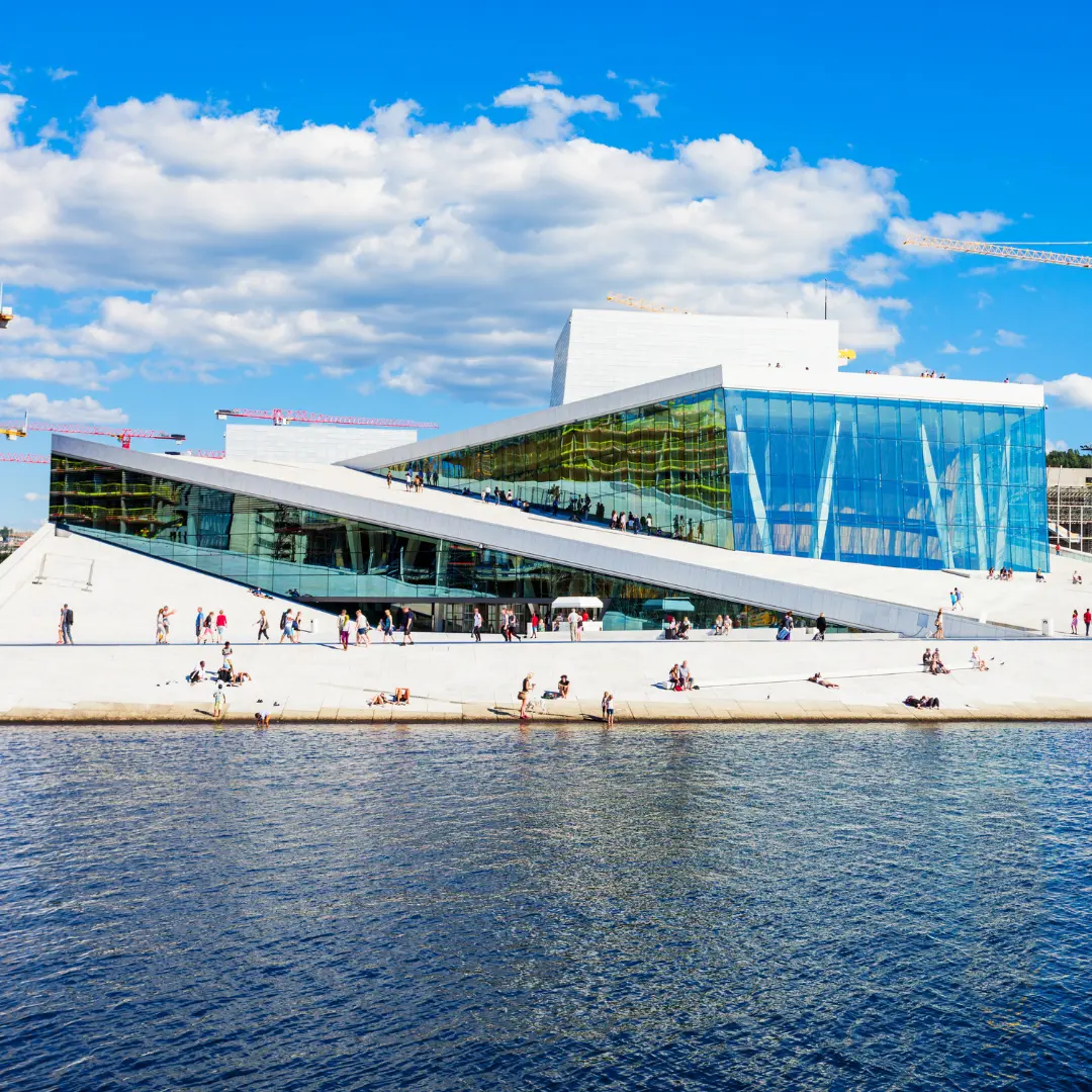 The Oslo Opera House, a modern angular glass and white stone building, sits beside the water under a blue sky with clouds. People walk and relax on the sloped roof and along the waterfront—contact F24 for more information.