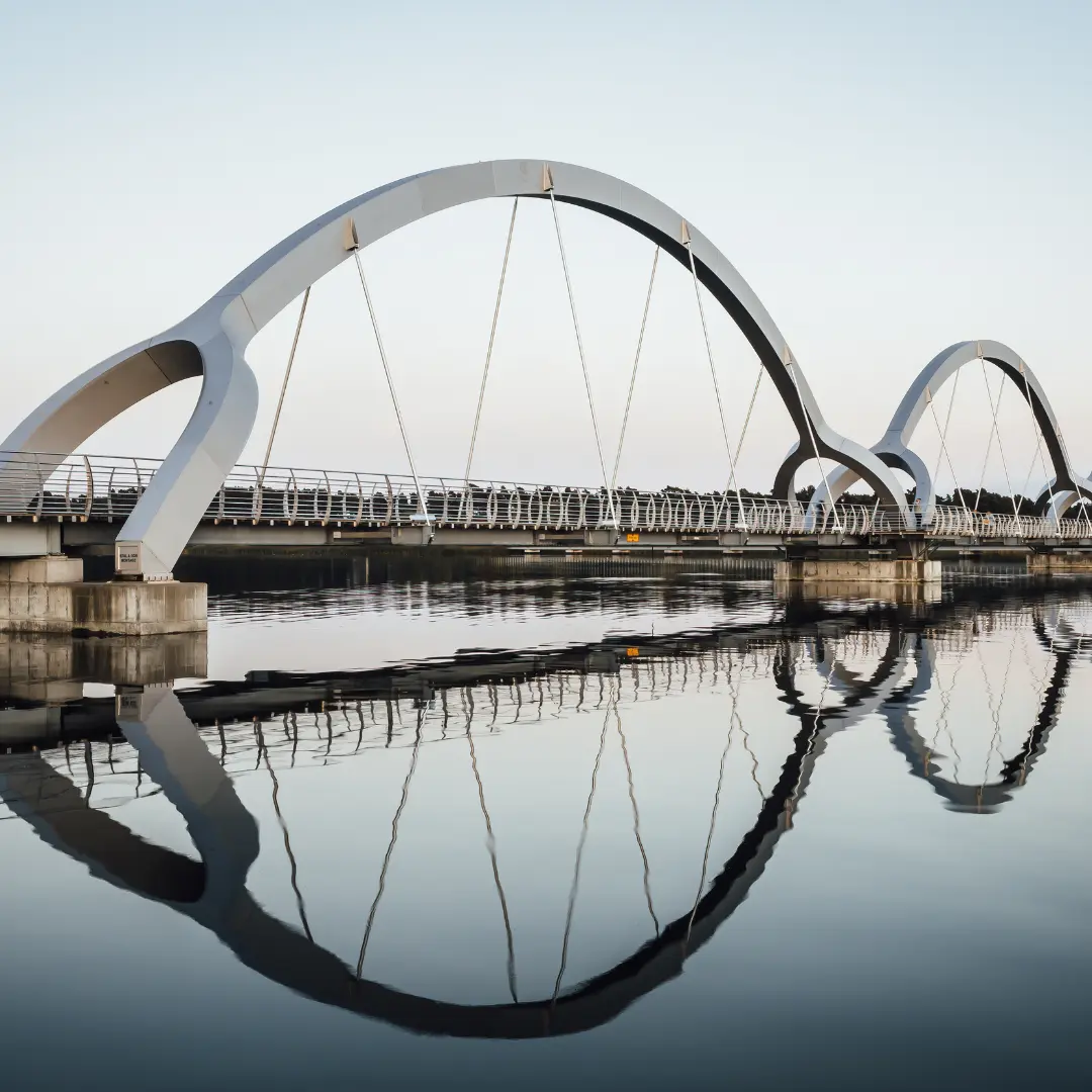 A modern arched pedestrian bridge with curved supports is reflected in calm water below, creating a contact f24 mirror-like effect. The clear, light blue sky enhances the tranquil atmosphere and perfect symmetry.