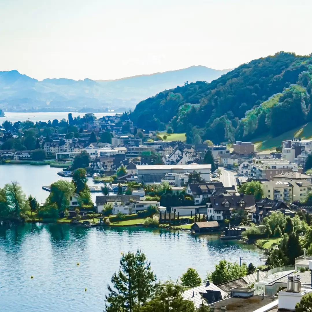 A scenic lakeside town with clusters of buildings, lush green hills, and trees. The calm lake reflects the landscape, distant mountains are visible under a clear sky. For more details or inquiries, contact f24.