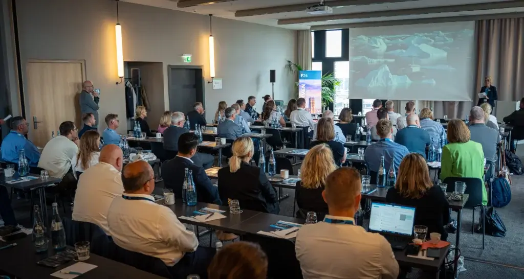 Die Konferenzteilnehmer sitzen an Tischen und blicken auf einen Referenten, der eine Präsentation über Eisberge hält. Viele machen sich Notizen oder arbeiten am Laptop und nutzen die Gelegenheit, sich während der gesamten Veranstaltung auszutauschen und Kontakte zu knüpfen.