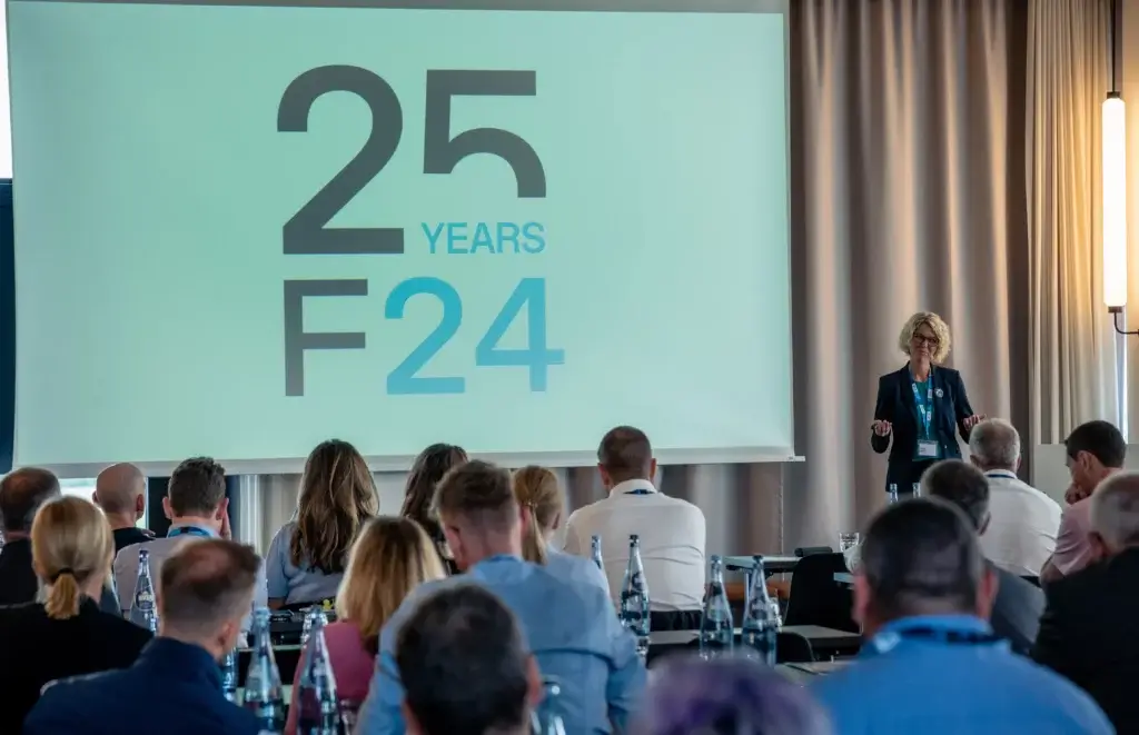 Eine Frau spricht vor Publikum in einem Konferenzraum und schafft dabei eine Atmosphäre, die alle dazu anregt, sich auszutauschen und Kontakte zu knüpfen. Auf einer großen Leinwand ist in fetter Schrift „25 YEARS F24“ zu lesen, während auf den Tischen Flaschen und Notizblöcke bereitstehen.