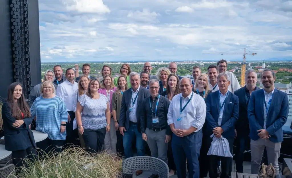 Eine Gruppe von etwa 28 Personen posiert gemeinsam im Freien auf einem Dach, gekleidet in Business-Casual-Kleidung, vor dem Hintergrund einer Stadtlandschaft und eines blauen Himmels.