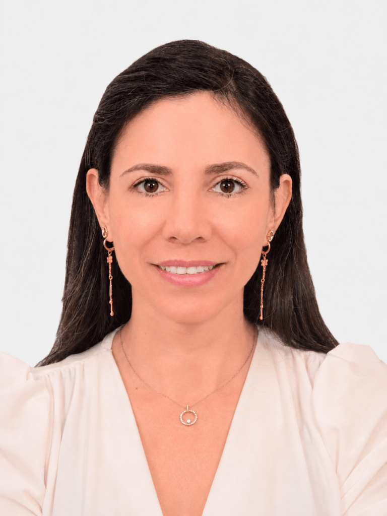 Woman with long dark hair, wearing gold earrings and a necklace, smiles slightly. She is dressed in a white blouse, and the background is plain and light-colored, reflecting un aire de profesionalismo y capacidades organizacionales.