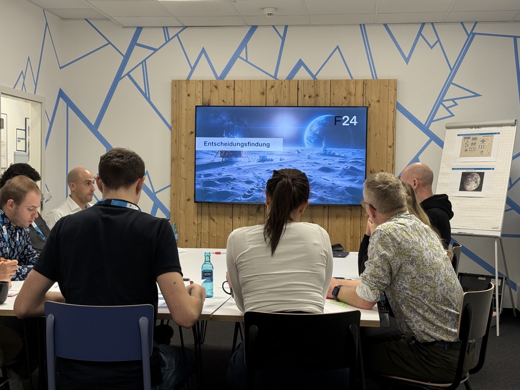 Eine Gruppe an Menschen die in einem Meeting Raum sitzen und auf einen Bildschirm schauen.